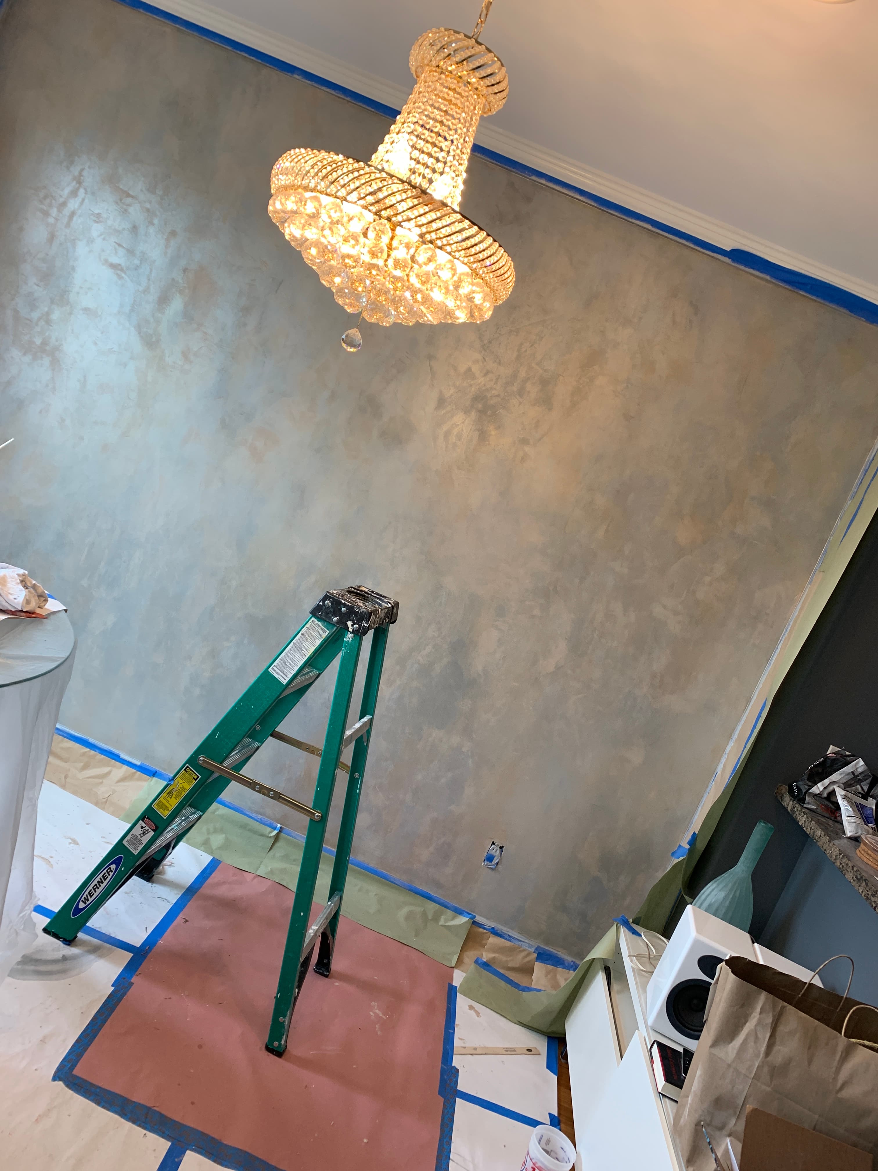Green stepladder and glowing crystal chandelier against a newly textured metallic grey accent wall.