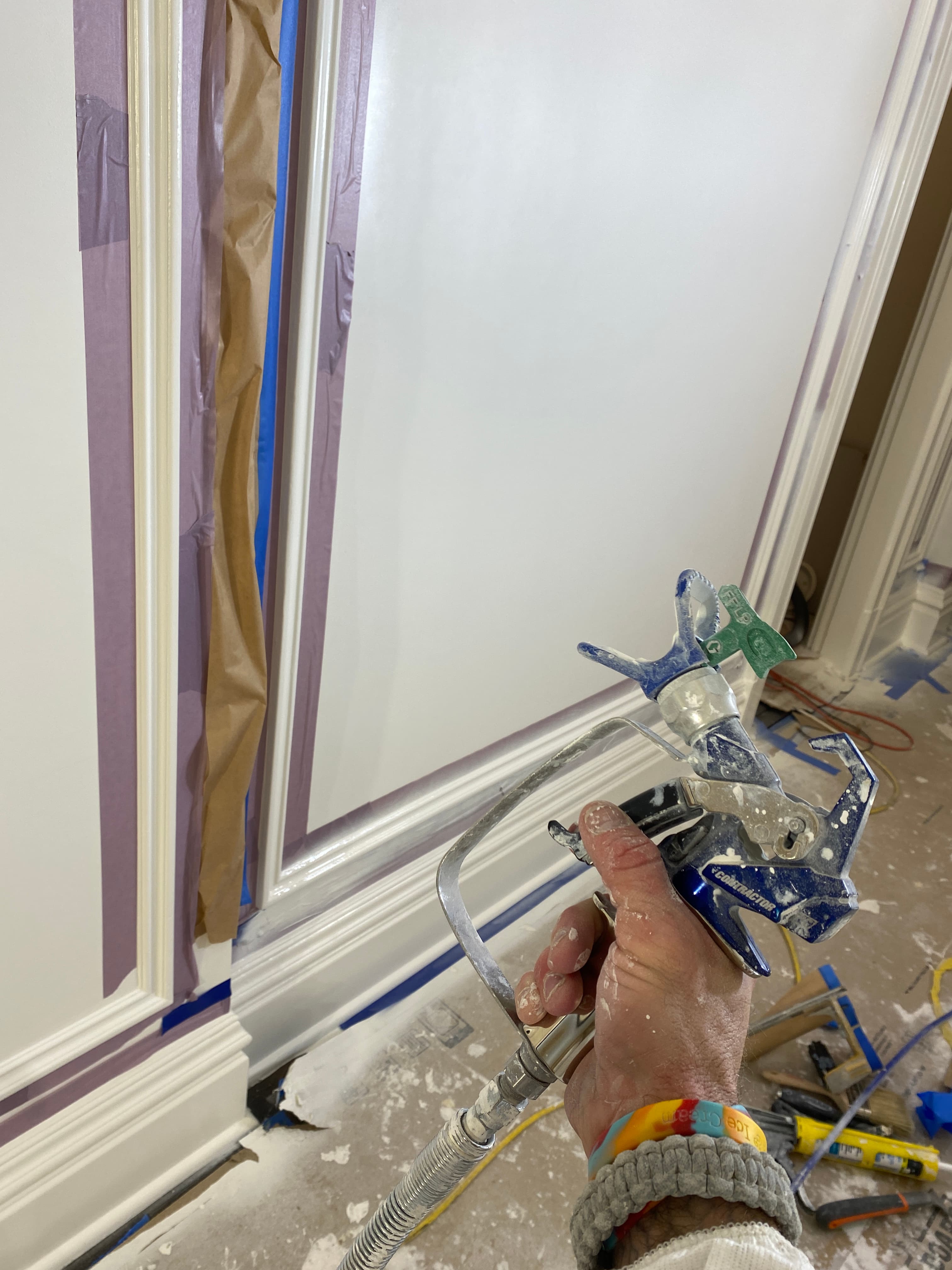 Hand holding a paint sprayer near white trim masked with purple and blue painter's tape.