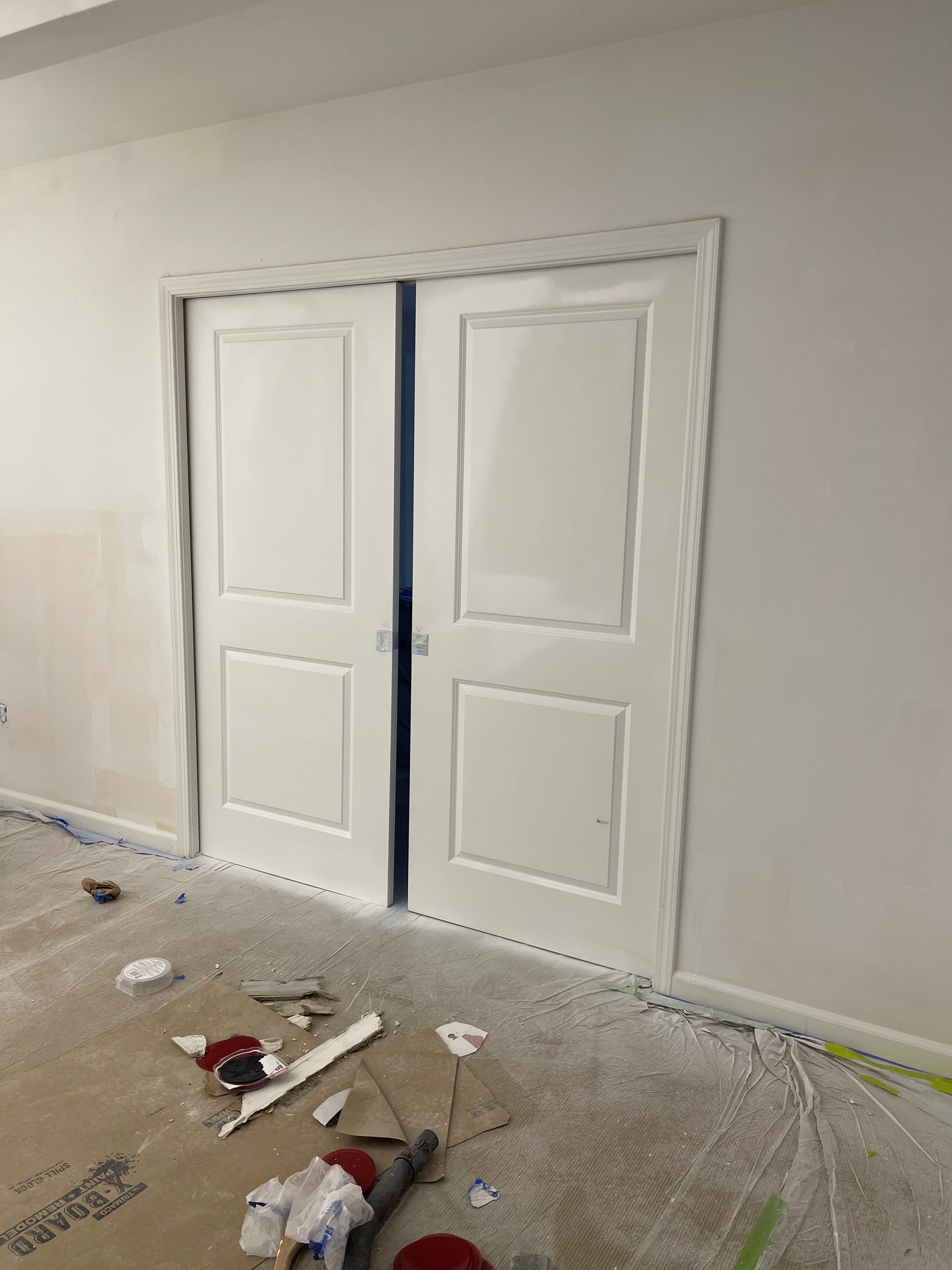 White double doors slightly open in a room with plastic floor coverings and renovation debris.