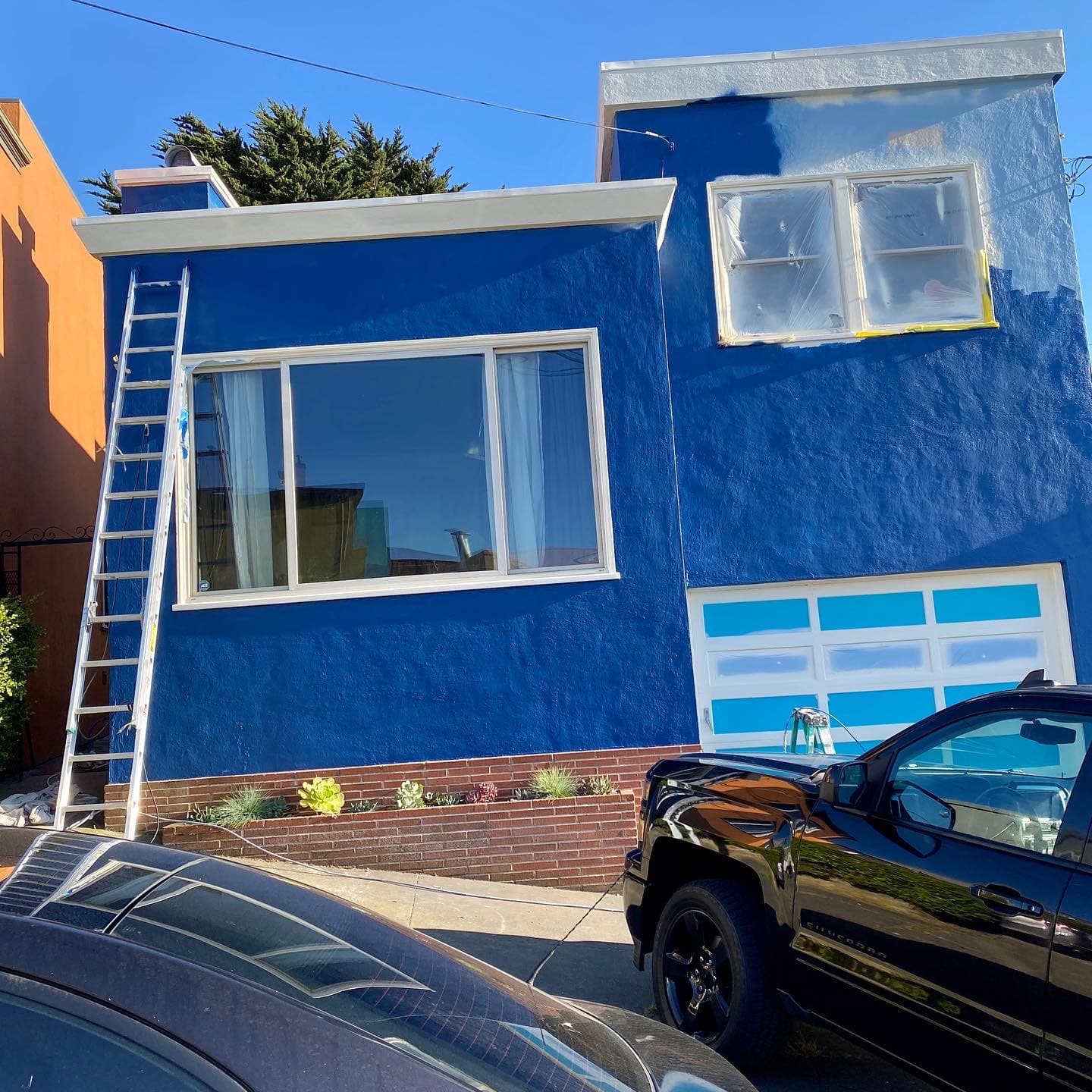 A house exterior being painted vibrant blue with a ladder and taped windows under sunlight.