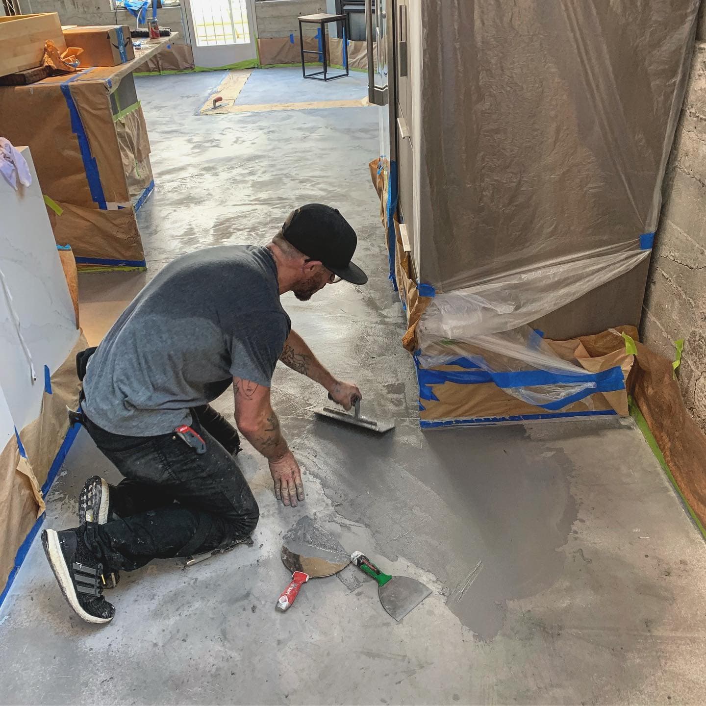 Man kneeling on a concrete floor, spreading grey resurfacing compound with a hand trowel.