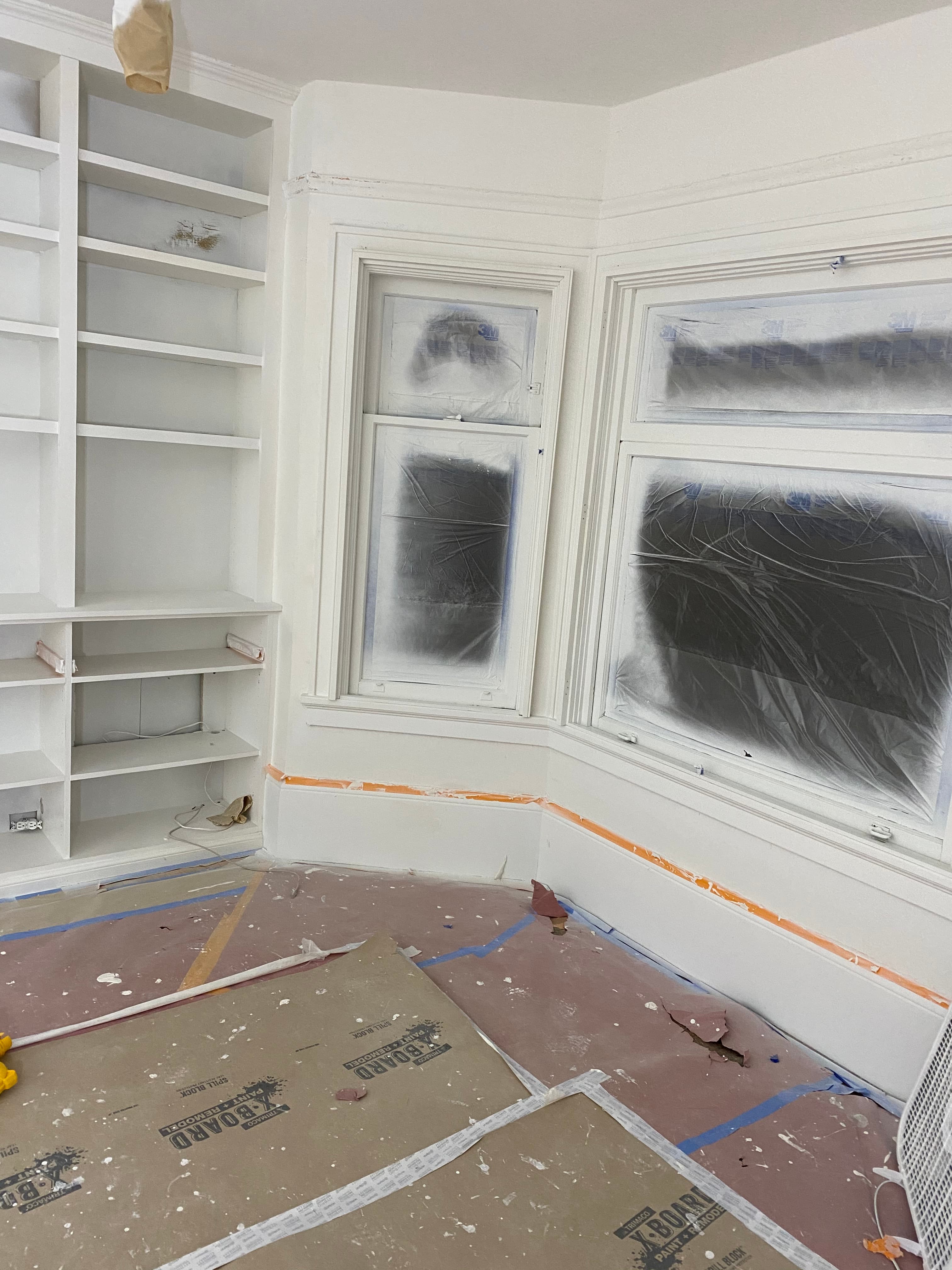 Interior room renovation featuring white built-in bookshelves and windows covered in protective plastic sheeting.