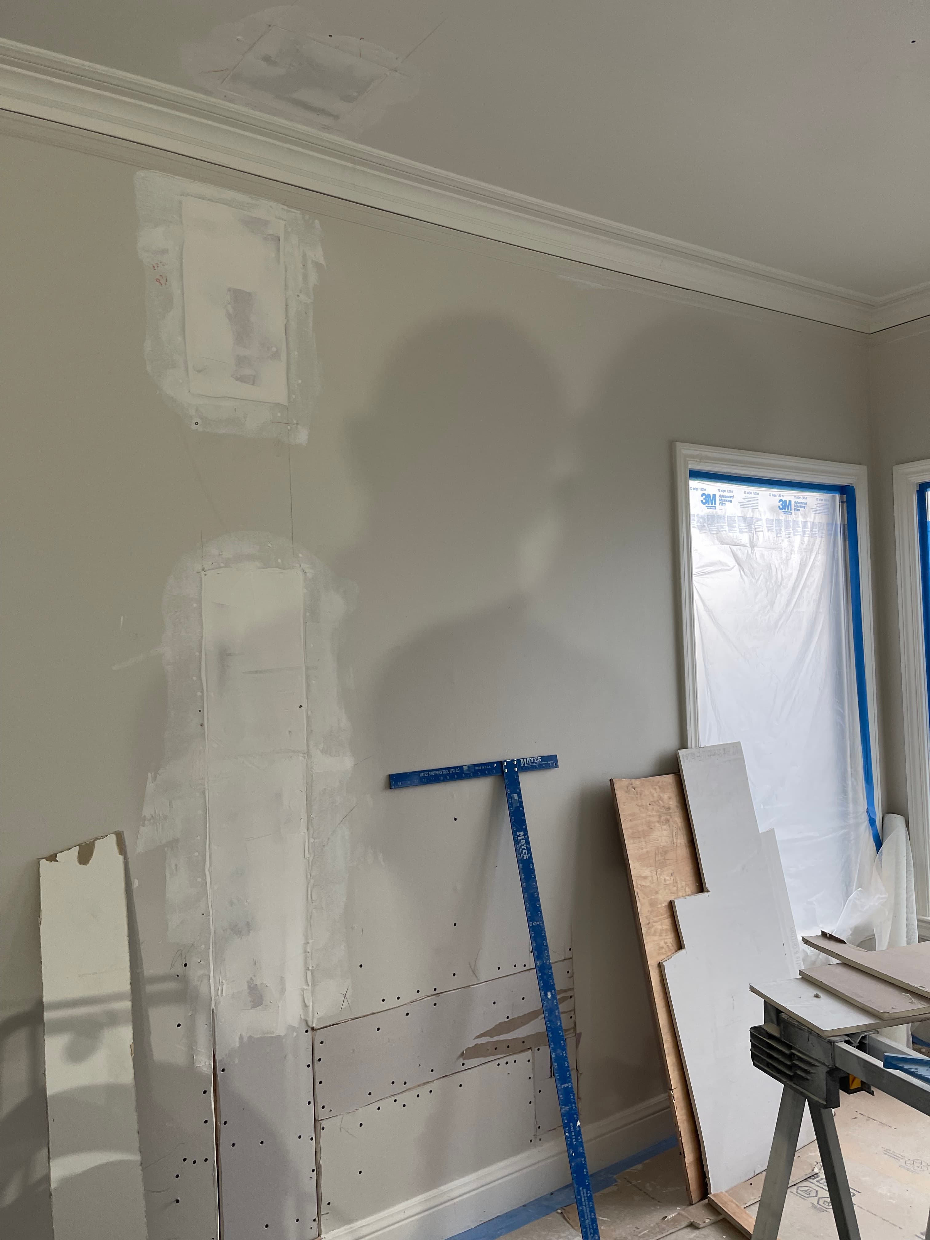 Patched drywall with fresh joint compound, a blue T-square, and a plastic-covered window during renovation.