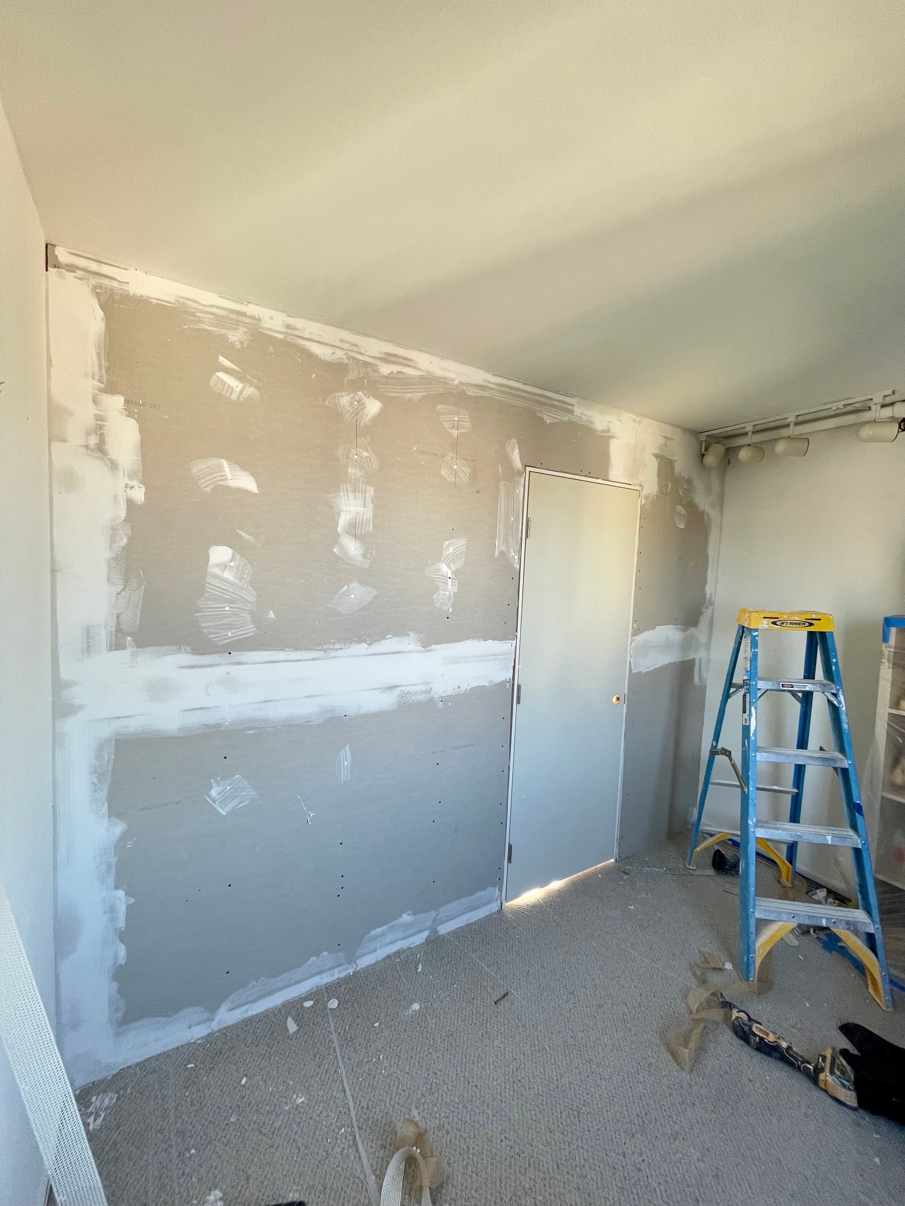Drywall wall with fresh joint compound on seams and screw holes beside a blue stepladder.