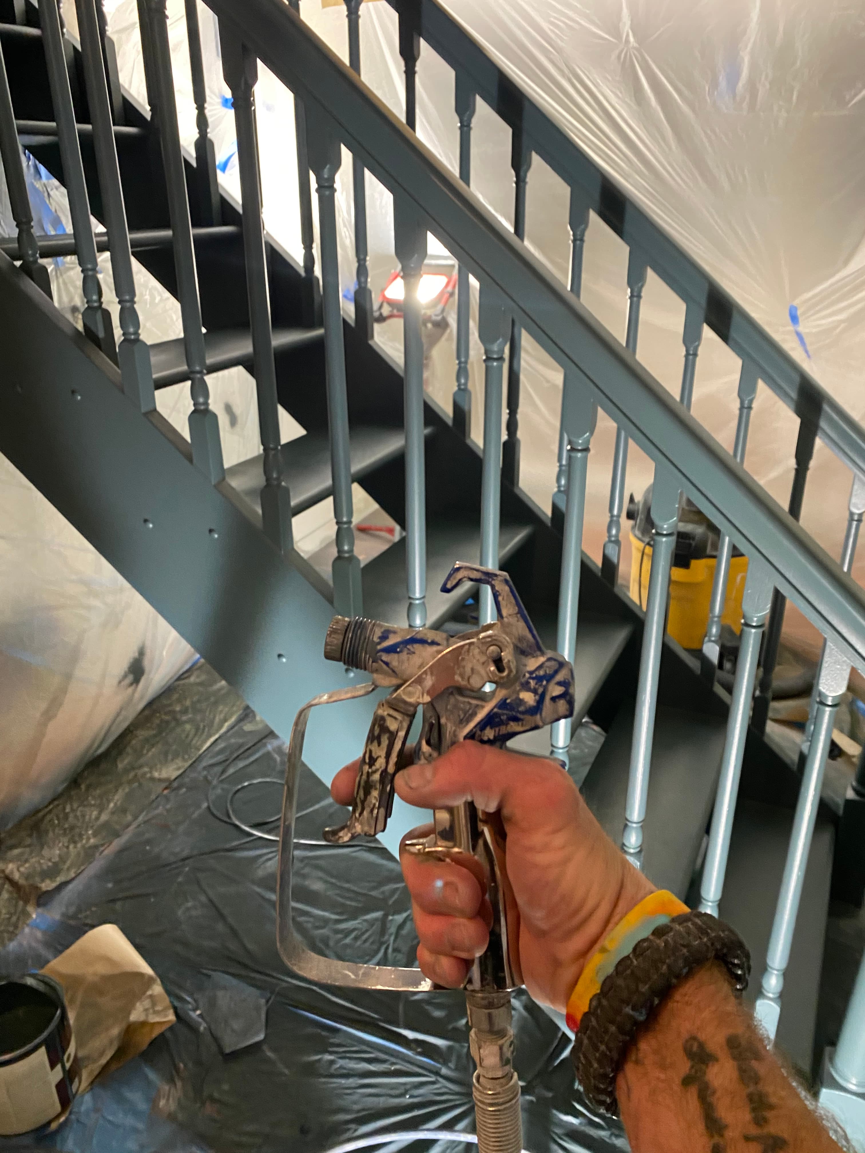 Hand holding a paint sprayer before a dark grey staircase protected by plastic sheeting.