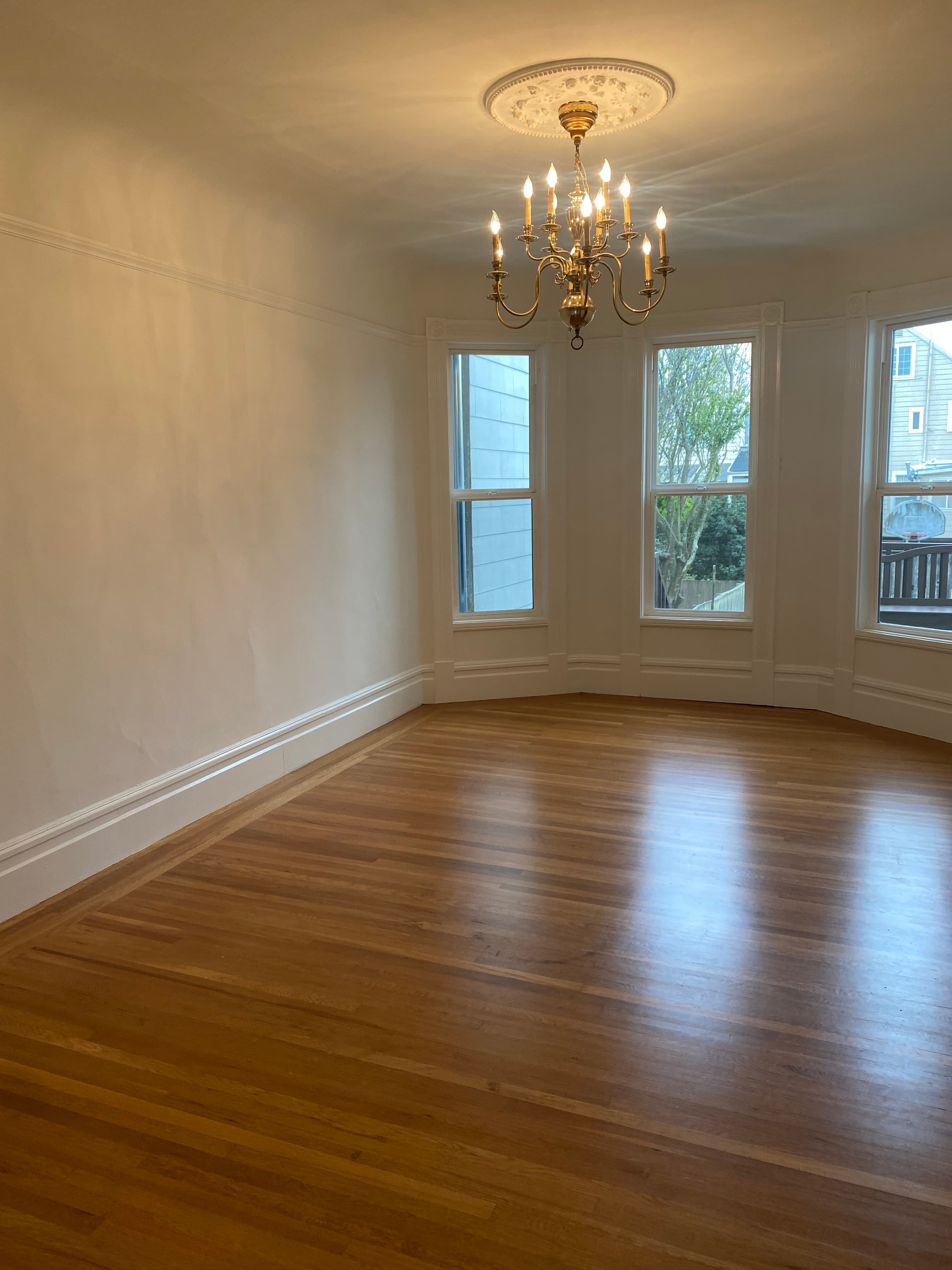 Empty room with gleaming hardwood floors, white walls, and an elegant brass chandelier hanging overhead.