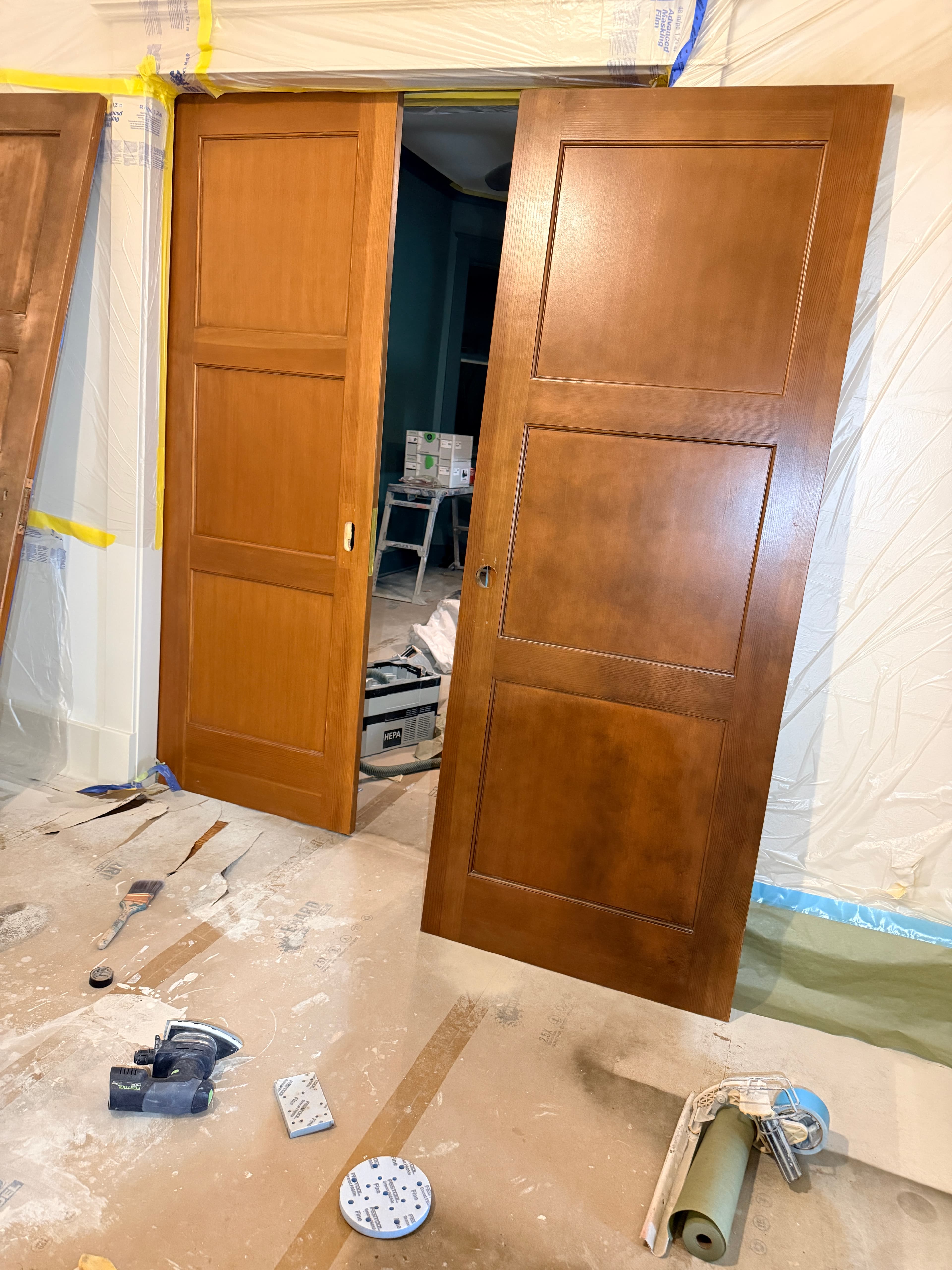 Two stained wooden doors leaning against plastic-covered walls with sanding tools on the floor.