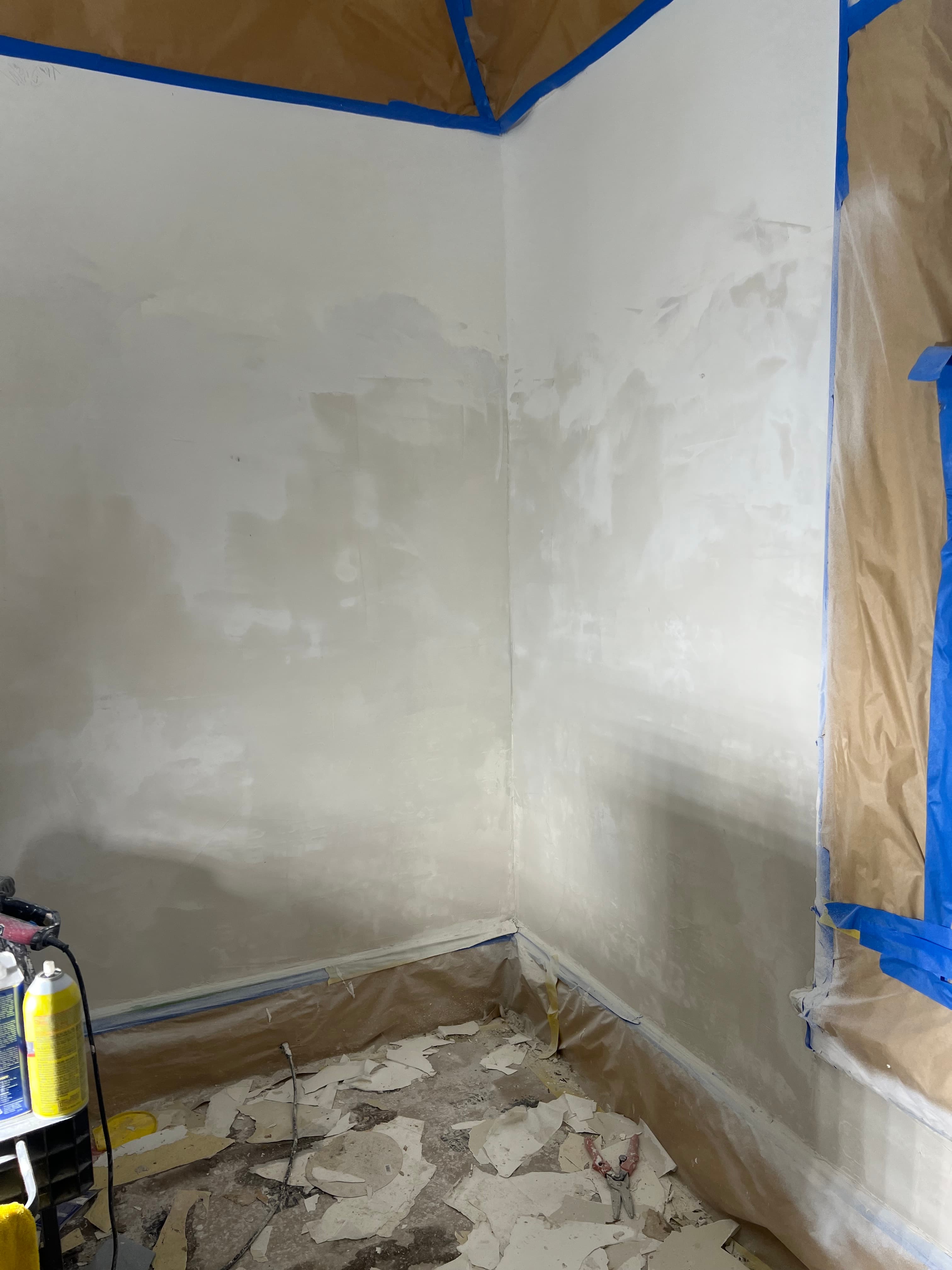Room corner under renovation with white plastered walls, blue painter's tape, and floor debris.
