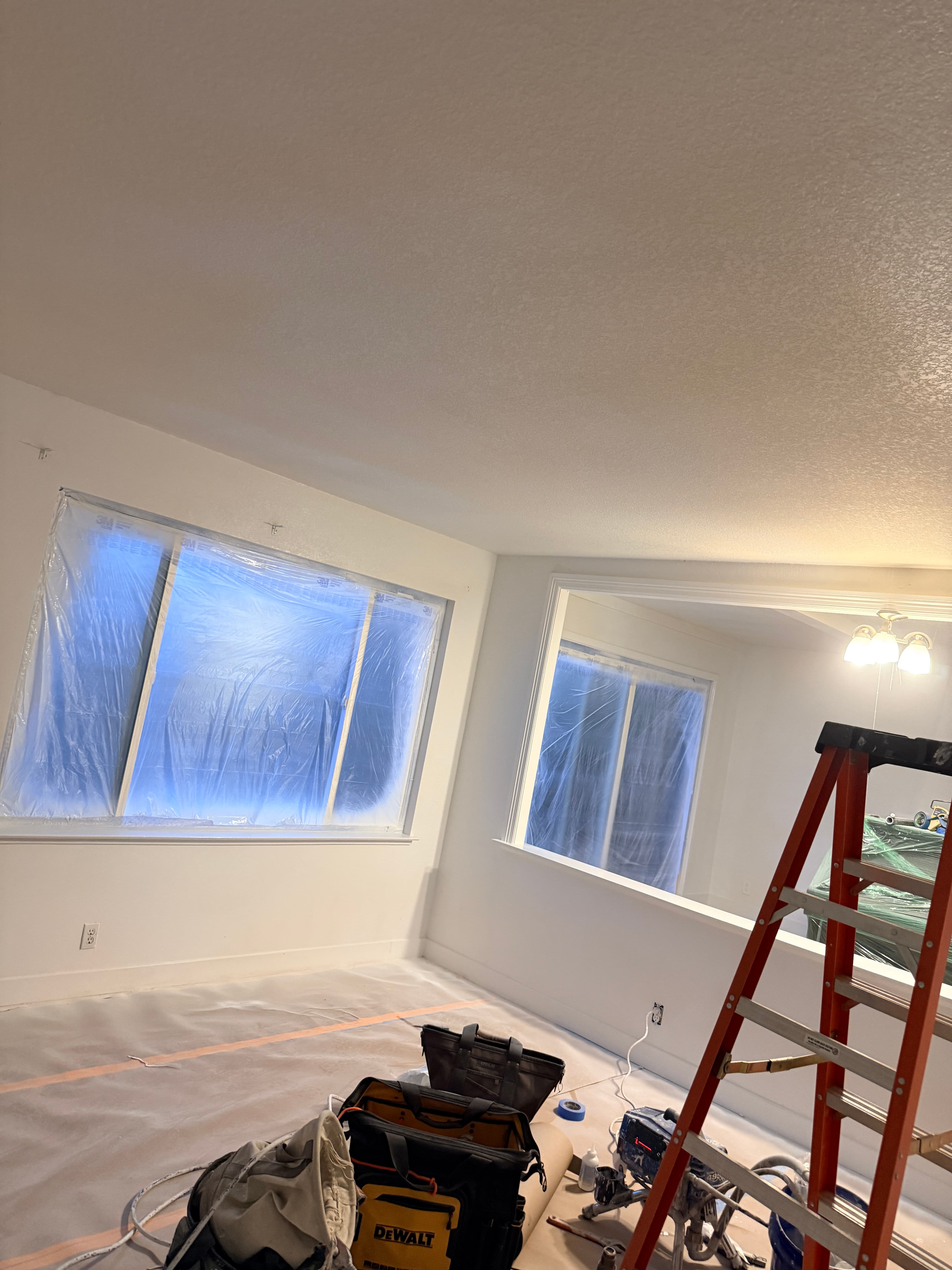 Interior room under renovation with blue plastic-covered windows, an orange ladder, and tool bags.