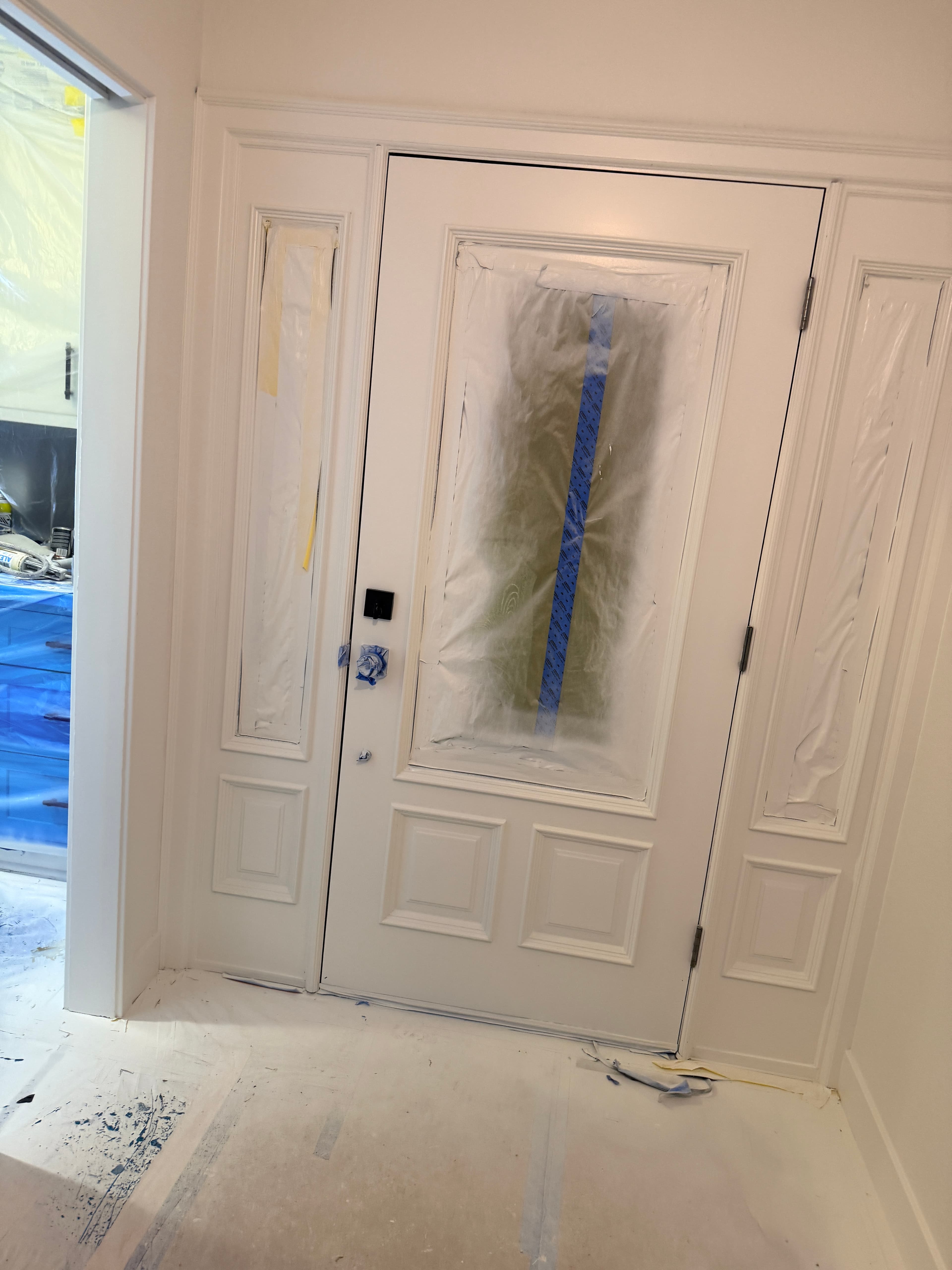 White front door with sidelights, glass panels masked with plastic and blue tape for painting.