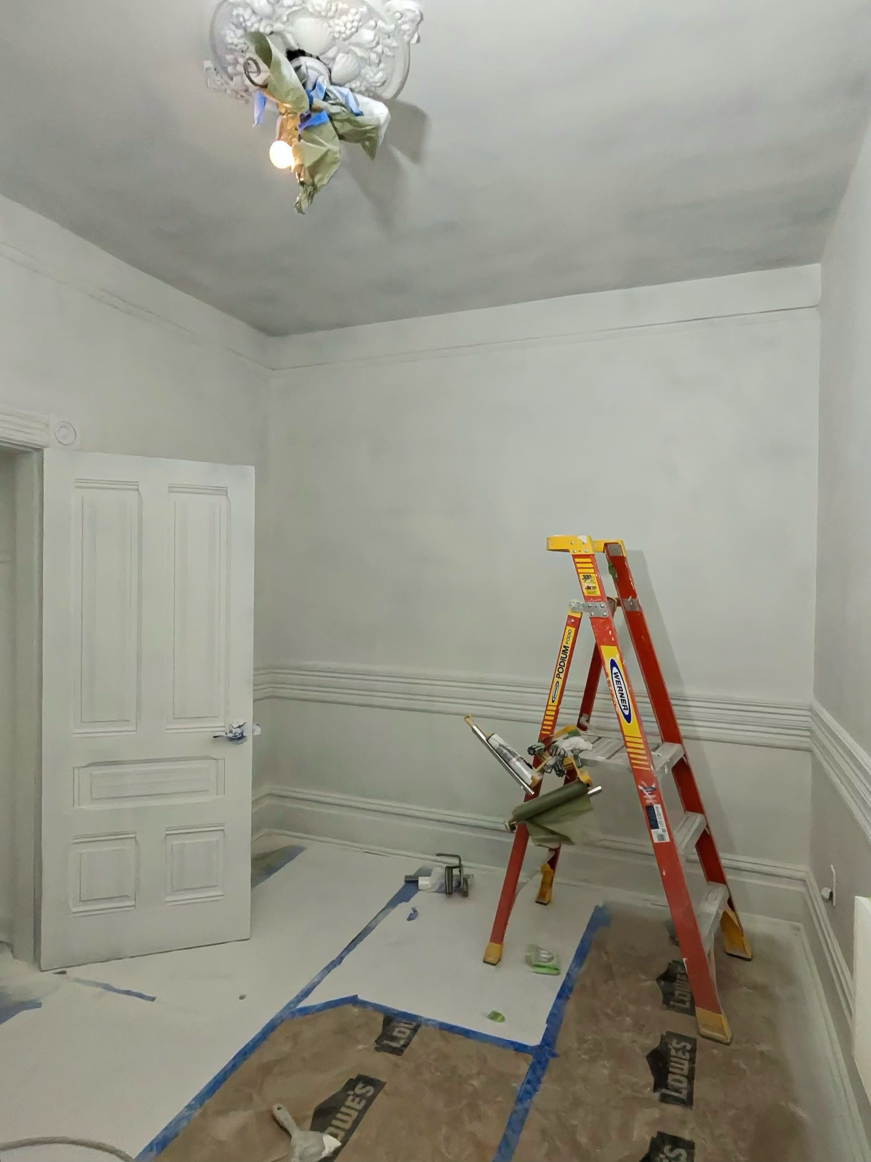 White room being painted with an orange ladder, floor protection, and an ornate ceiling medallion.